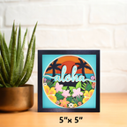 Framed artwork with 'aloha' text and tropical design on a wooden surface with a plant.
