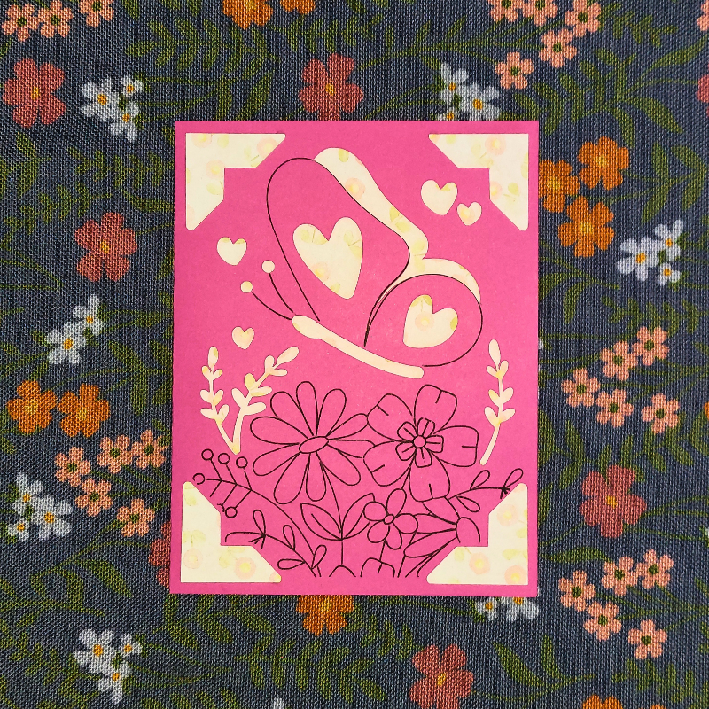 Decorative card with floral and heart designs on a pink background, placed on a fabric with floral patterns.