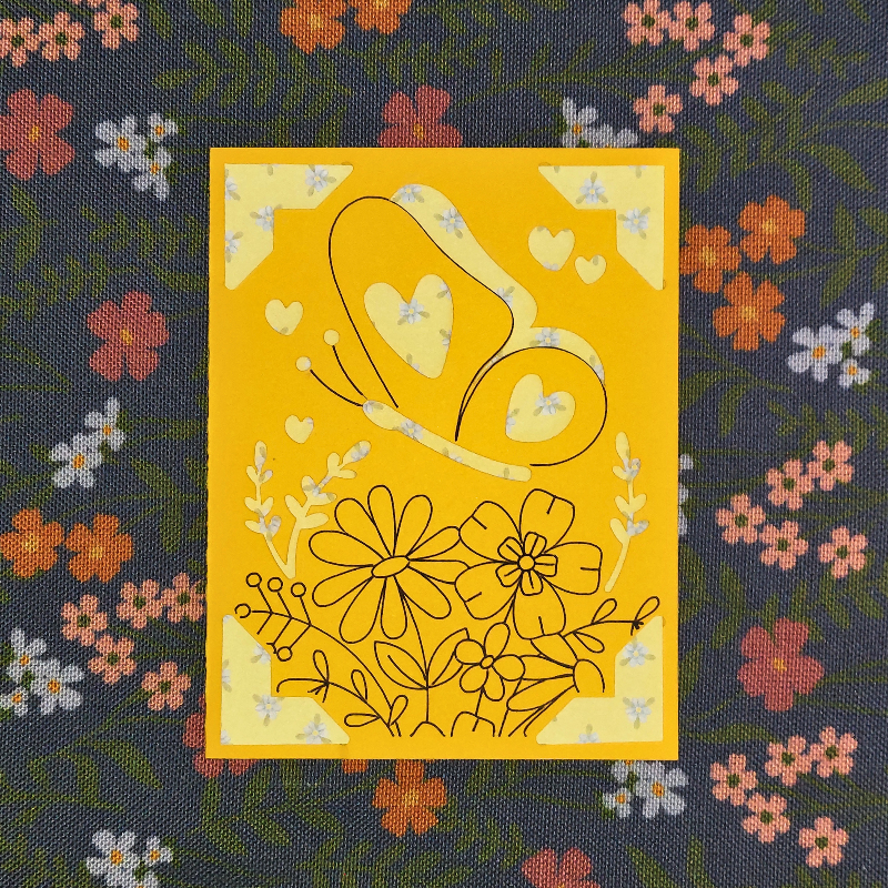 Yellow card with floral and heart designs on a dark fabric background with flowers.
