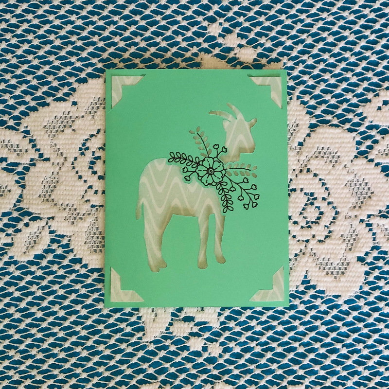 Card with a goat illustration on a green background, placed on a textured fabric surface.
