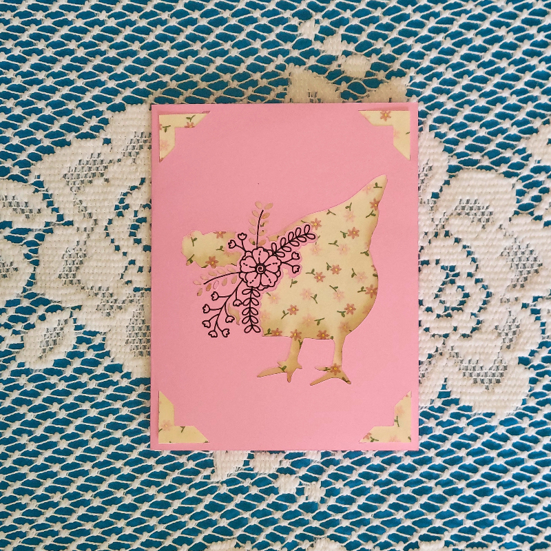 Card with floral chicken design on a pink background, placed on a textured fabric surface.