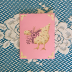 Card with floral chicken design on a pink background, placed on a textured fabric surface.