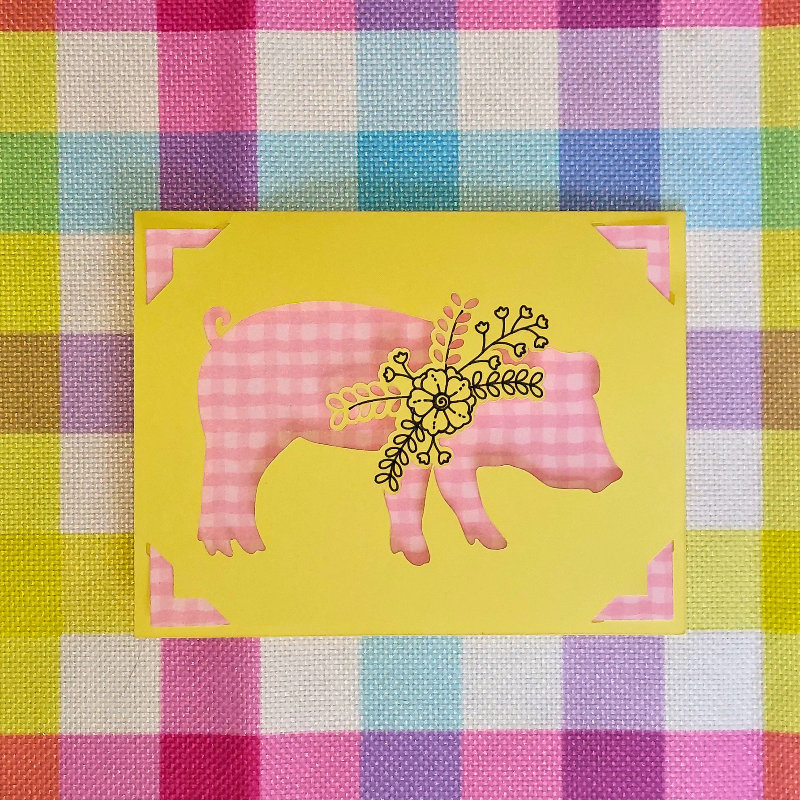 Pink pig design on a yellow card with decorative elements, placed on a colorful checkered fabric background.