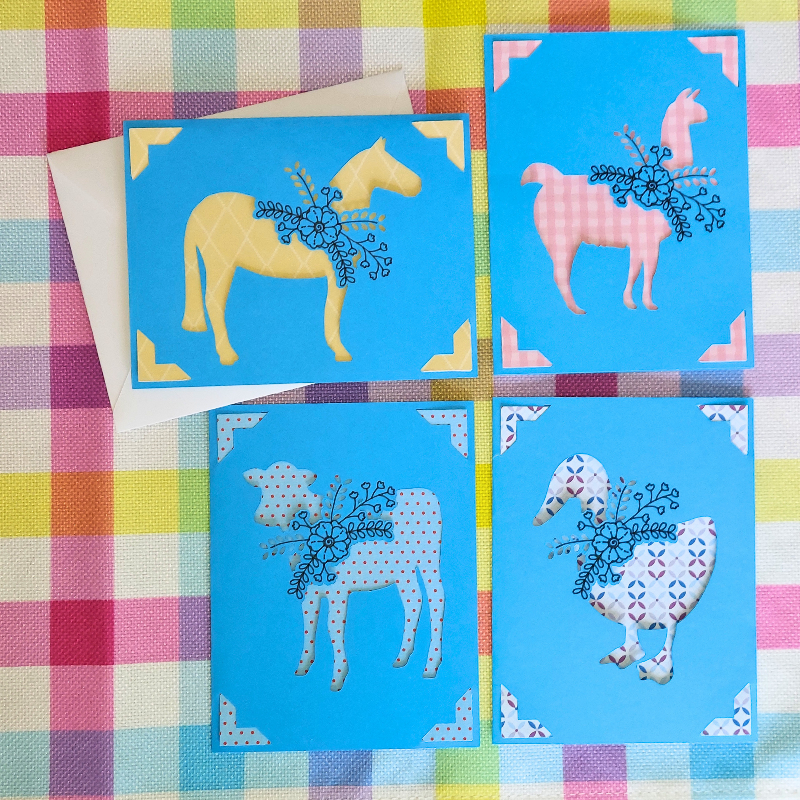 Four colorful greeting cards with animal illustrations on a checkered fabric background.