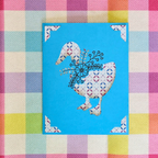 Decorative card with a goose design on a blue background, placed on a colorful checkered fabric.