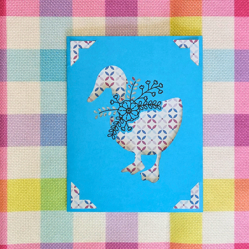 Decorative card with a goose design on a blue background, placed on a colorful checkered fabric.