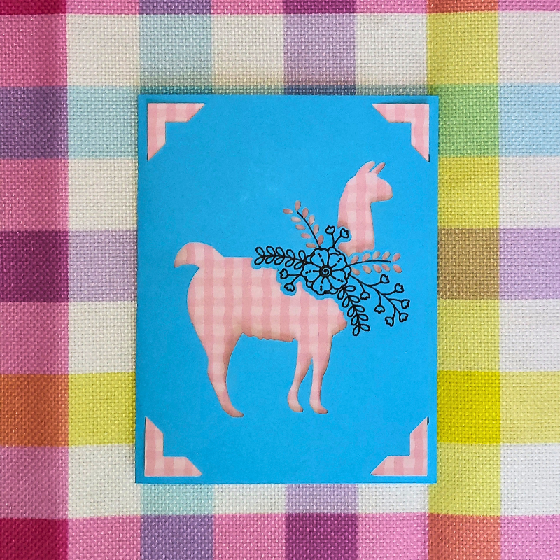 Card with pink llama design on blue background, placed on a checkered fabric background.