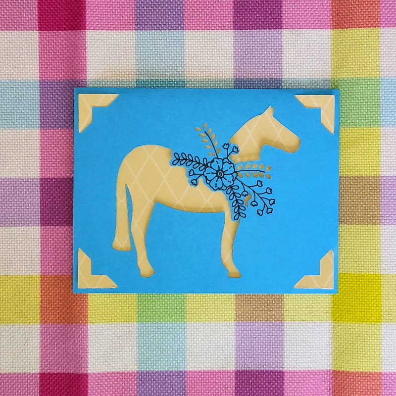 Decorative card with a horse and floral design on a blue background, placed on a checkered fabric.