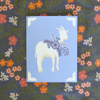 Decorative card with a white goat illustration on a blue background, placed on a floral fabric background.