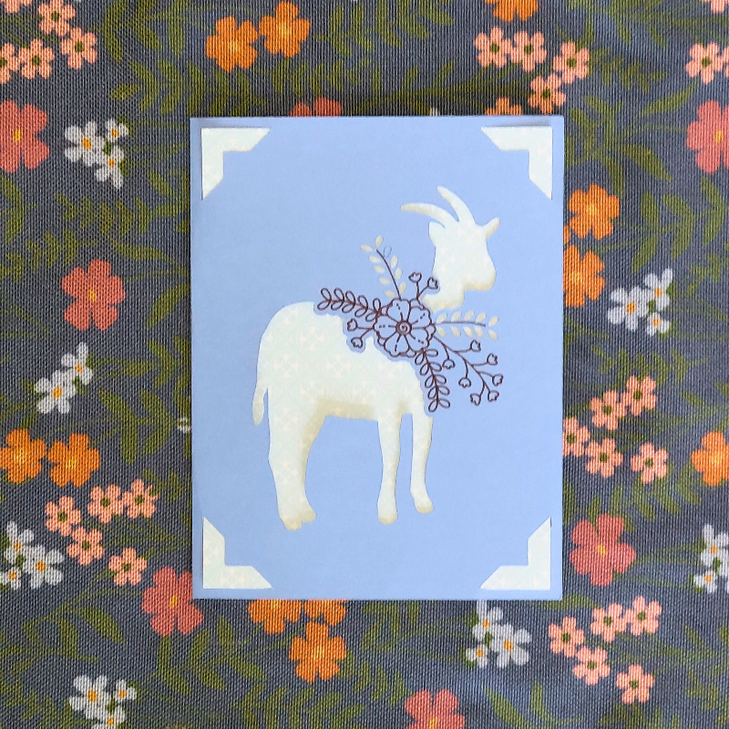 Decorative card with a white goat illustration on a blue background, placed on a floral fabric background.