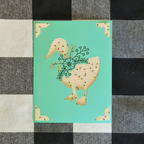 Card with a goose design on a turquoise background, placed on a checkered fabric surface.