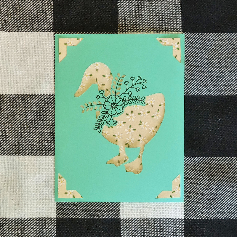Card with a goose design on a turquoise background, placed on a checkered fabric surface.