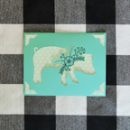 Card with a pig illustration on a turquoise background, placed on a checkered fabric surface.
