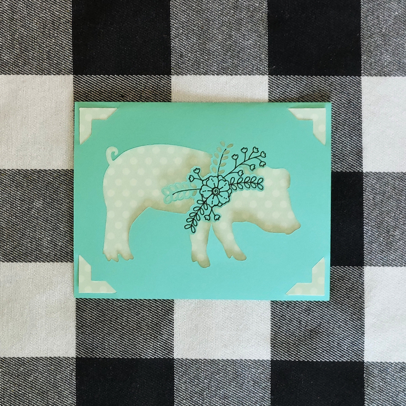 Card with a pig illustration on a turquoise background, placed on a checkered fabric surface.