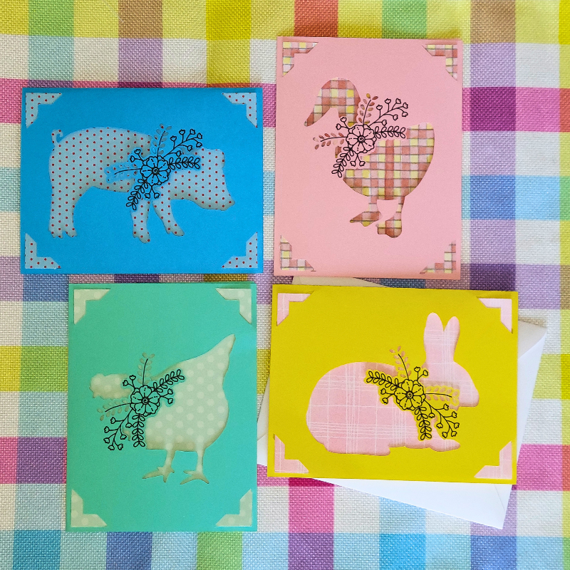 Four colorful greeting cards with animal illustrations on a checkered fabric background.