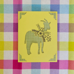 Decorative card with a goat design on a yellow background, placed on a colorful checkered fabric.