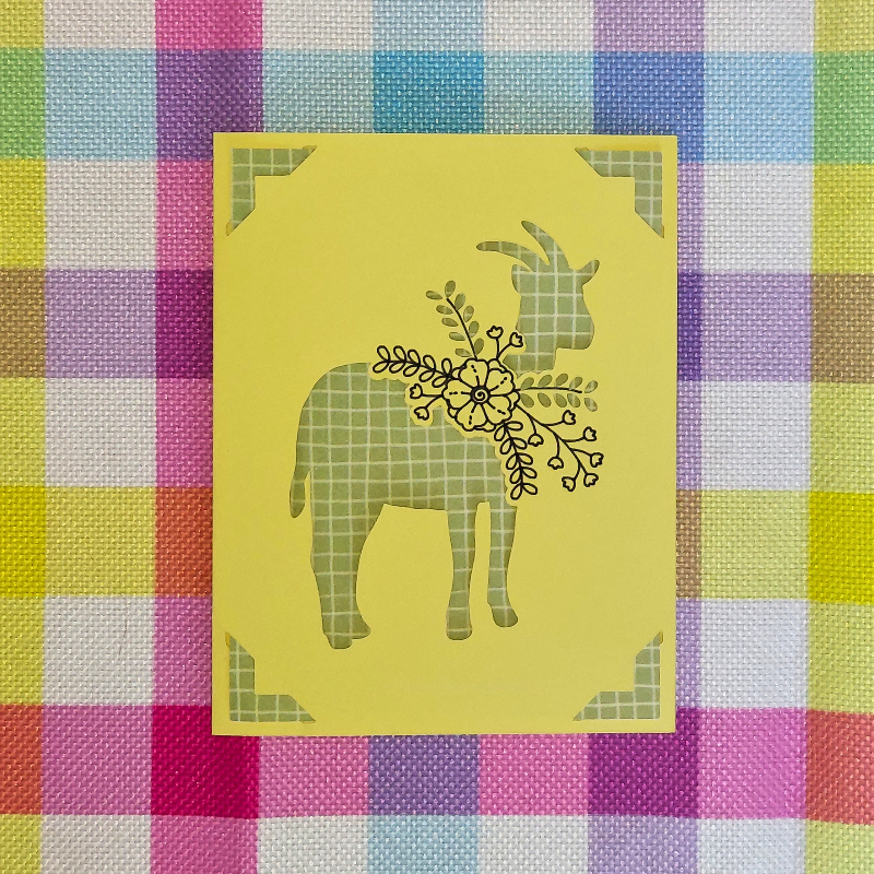 Decorative card with a goat design on a yellow background, placed on a colorful checkered fabric.