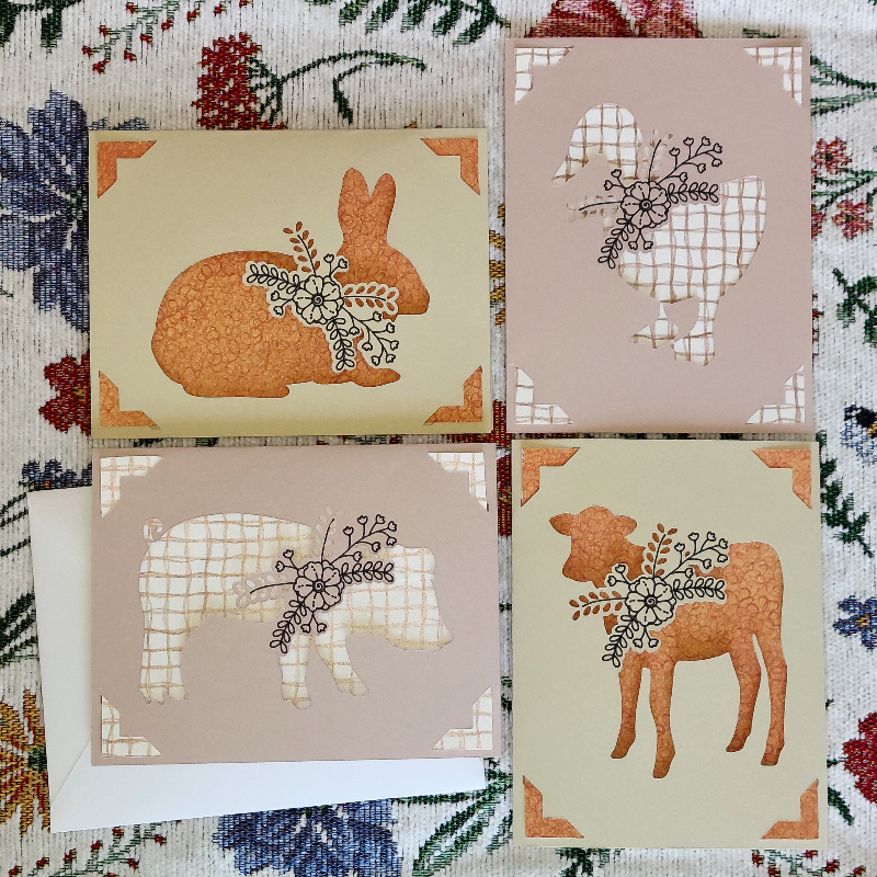Four greeting cards with animal silhouettes and floral designs on a patterned fabric background.
