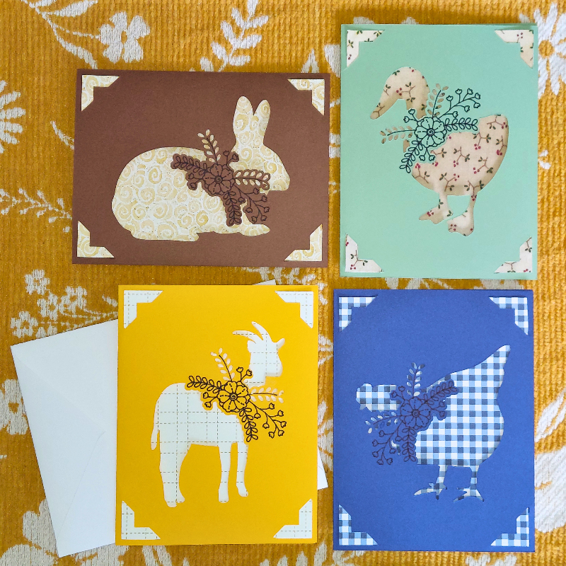 Four greeting cards with animal illustrations on a floral fabric background