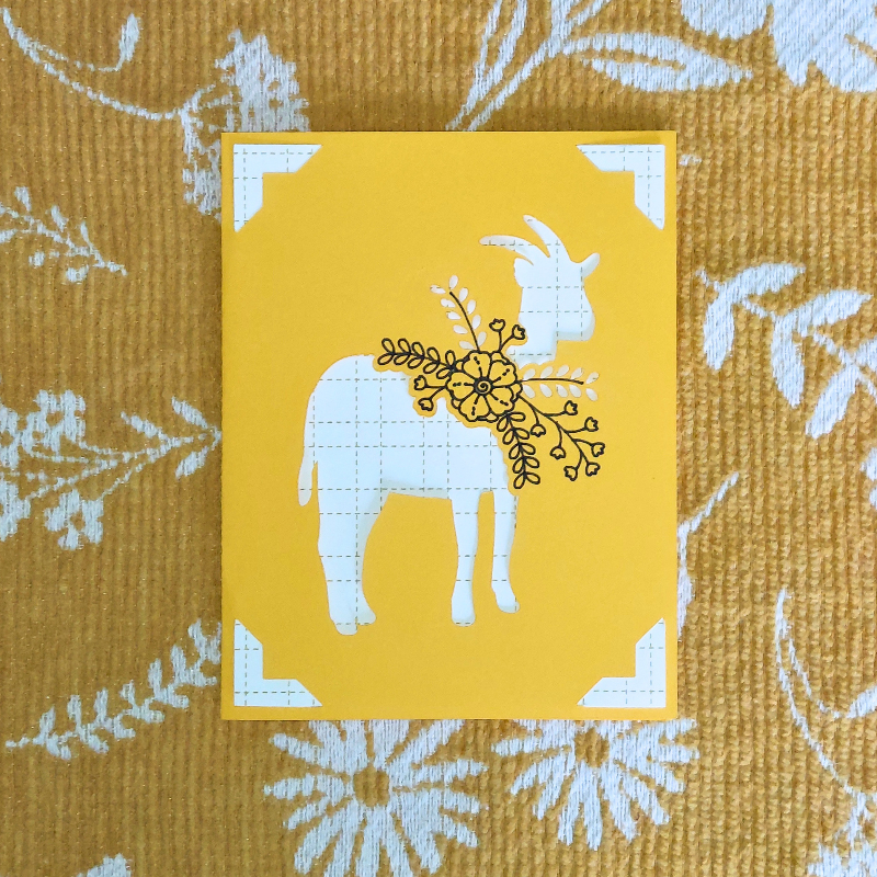 Card with white goat illustration on a yellow background, placed on a textured fabric surface.