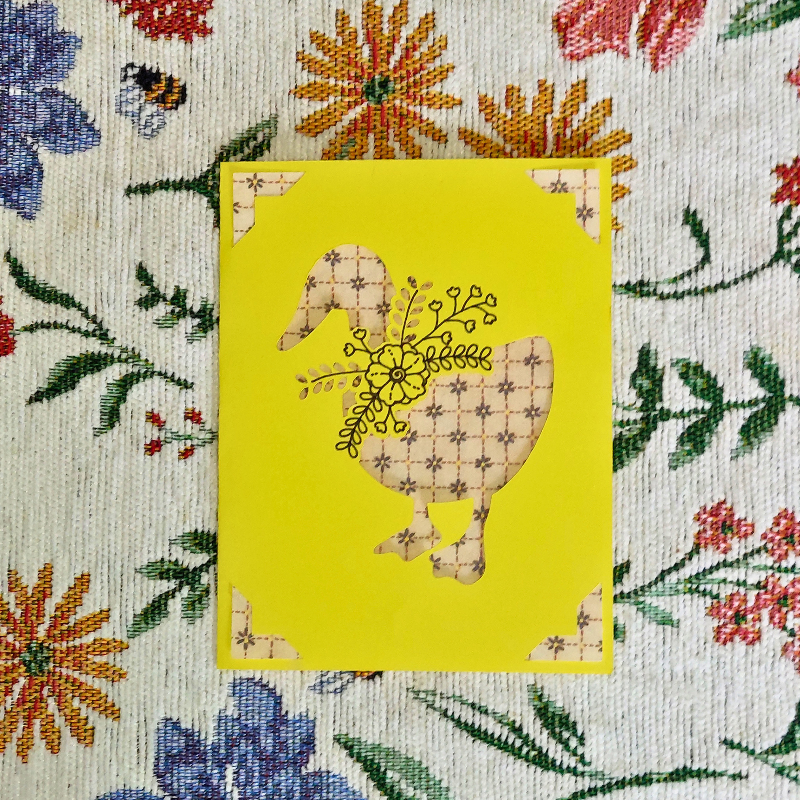 Decorative card with a goose design on a yellow background, placed on a floral fabric background.