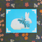 Fabric with a white rabbit holding a blue flower on a blue square, surrounded by floral patterns.