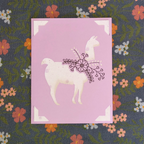 Card with a llama illustration on a purple background, featuring floral elements, on a fabric background with flowers.
