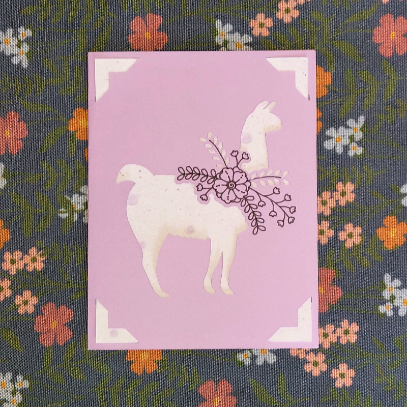 Card with a llama illustration on a purple background, featuring floral elements, on a fabric background with flowers.