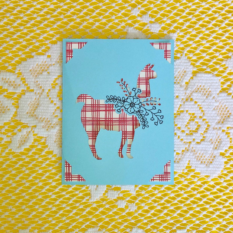 Card with a plaid llama design on a blue background, placed on a textured yellow and white surface.