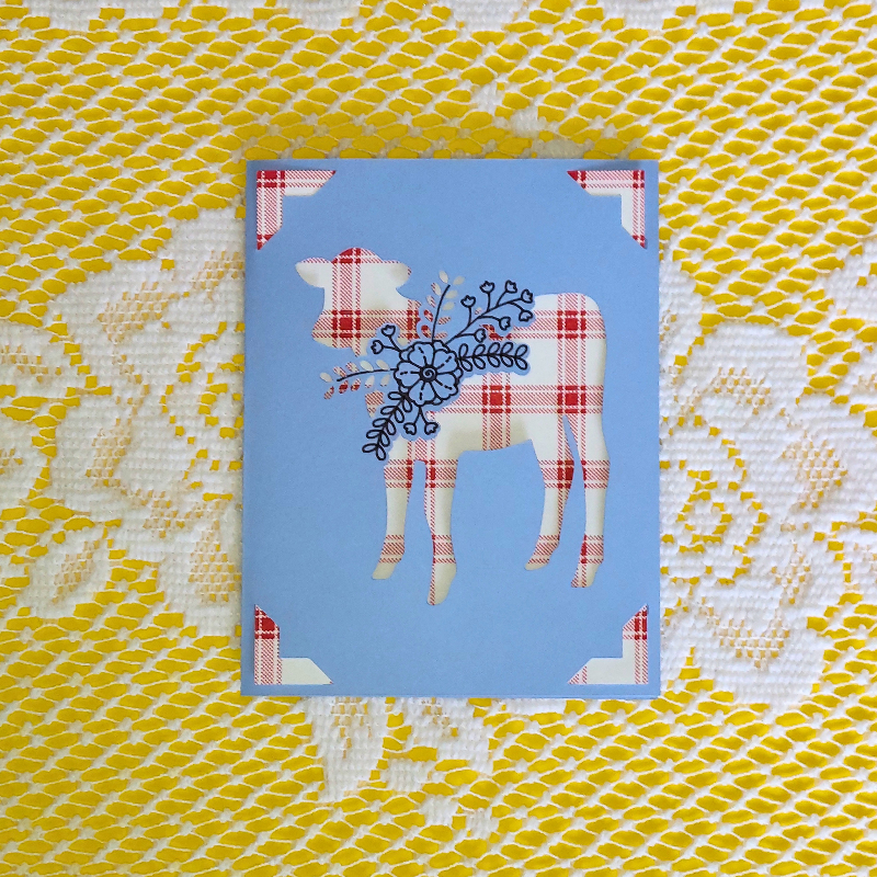 Decorative card with a plaid cow design on a blue background, placed on a yellow and white patterned fabric.
