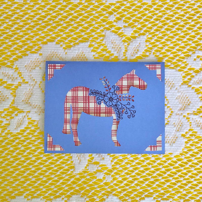 Card with red and white patterned horse design on a blue background, placed on a yellow and white textured fabric.