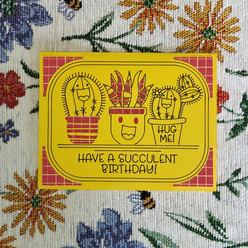 Cartoon succulents with a birthday message on a yellow background, placed on a floral fabric.