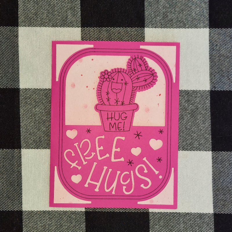 Pink card with a cactus illustration and 'Hug Me! Free Hugs!' text on a checkered fabric background.
