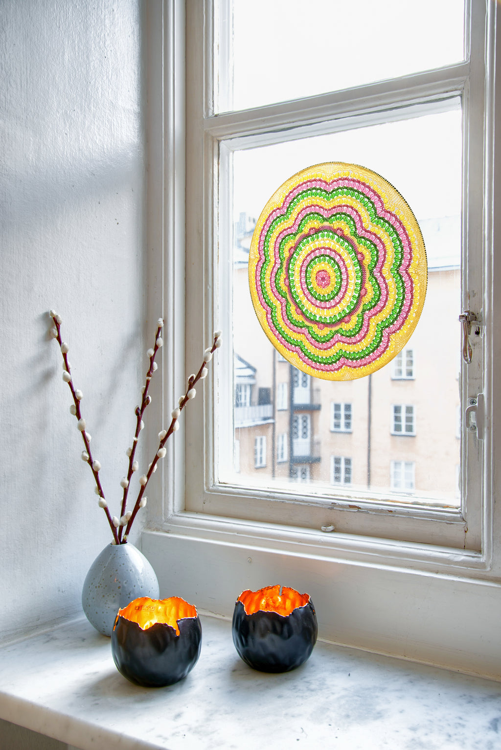 Decorative mandala on a window, two black candle holders with orange candles on a windowsill.