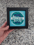 Framed decorative piece with 'Nelson Family' design held by a hand on a granite surface