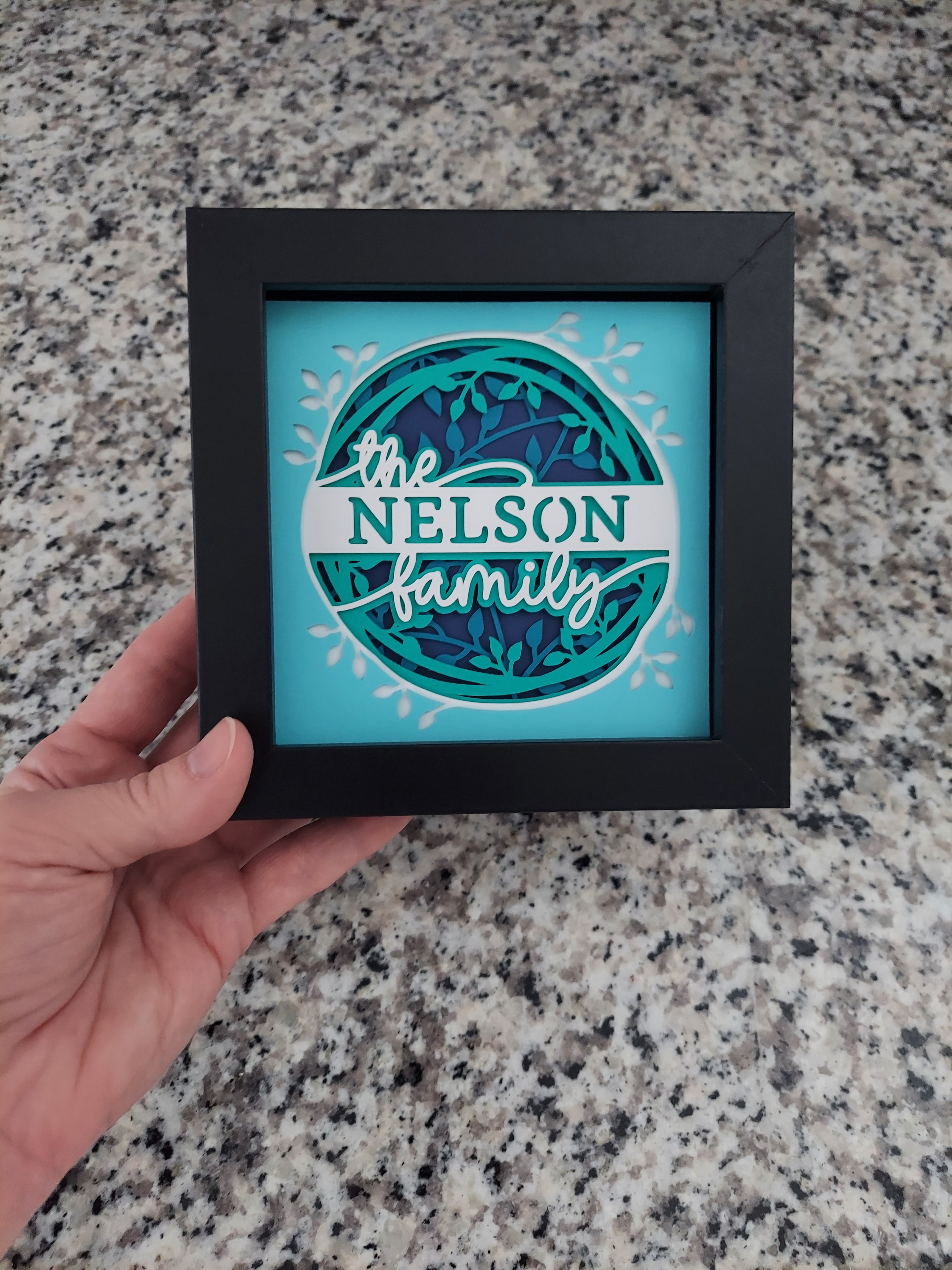 Framed decorative piece with 'Nelson Family' design held by a hand on a granite surface